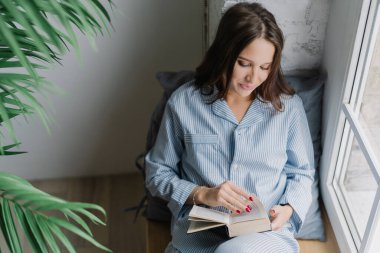 Üstten görünüm kitap haline duruldu karanlık uzun saçlı memnun kadın heyecan verici bir şey okur, rahat bir atmosfer evde sahiptir, pencere pervazına boş odada teşkil etmektedir. İçin zaman okuma ve dinlenme.