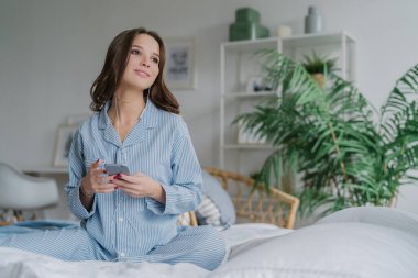 Shot oldukça esmer öğrenci dil yolu ile mobil app öğrenir, ses kulaklık ile dinler, serbest zaman var, gündelik kıyafeti giymiş, rahat beyaz yatakta teşkil etmektedir, bir kenara düşünceli bir görünüme sahip