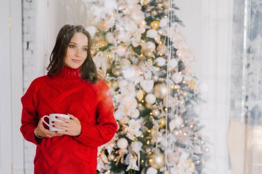 Düşünceli hoş örgü kırmızı kazaklı kadın arıyor, tutar kupa çay veya kahve ile standları Noel ağacı, bir şey hakkında düşünüyor yakınındaki hoş, boş kopya alanı reklam için bir kenara