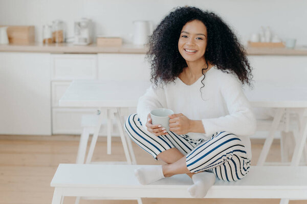 Счастливого утра. Фото веселой кудрявой афроамериканки, сидящей в позе лотоса на белой скамейке, пьющей вкусный ароматный напиток, расслабленной, позирующей против кухонного интерьера, широко улыбающейся
