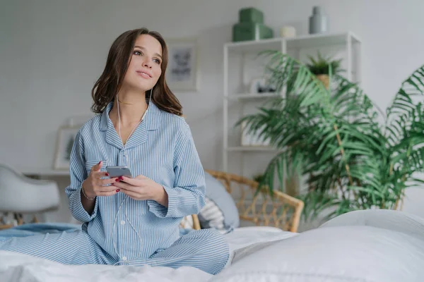 Shot oldukça esmer öğrenci dil yolu ile mobil app öğrenir, ses kulaklık ile dinler, serbest zaman var, gündelik kıyafeti giymiş, rahat beyaz yatakta teşkil etmektedir, bir kenara düşünceli bir görünüme sahip