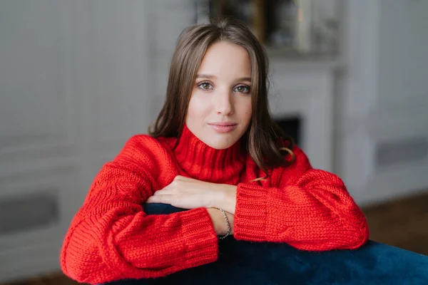 Calm pretty young Caucasian female dressed in warm knitted pullover, leans on armchair, looks with dark eyes directly at camera, poses against domestic interior indoor. People and beauty concept.