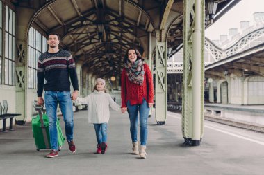 Shot of happy family going to have nice trip during holidays, carry bag, walk on railways station platform, being in good mood. Father, mother and child arrive from journey. Travelling concept