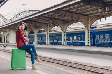 Horizontal shot of happy woman has telephone conversation, has spare time, sits at green bag, poses against railway station with copy space for your text. Communication and travelling concept