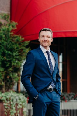 Vertical shot of attractive young male company owner celebrates something, stands outside, keeps hands in pocket, dressed in formal black suit, glad to have talk with someone, discuss work together