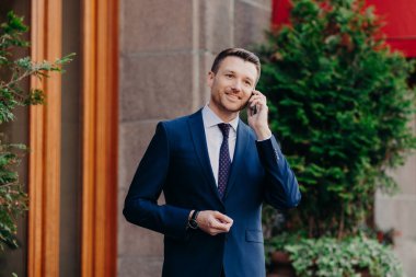 Successful businessman has work break, uses mobile phone for having talk, being always on line, dressed in formal suit, has pleasant smile on face, stands outdoor near restaurant. Technology concept