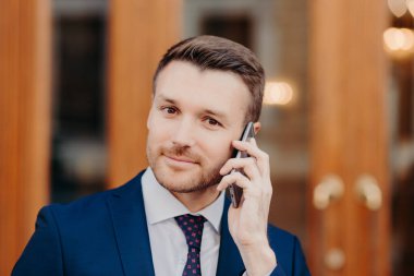 Close up portrait of good looking male with attractive appearance, speaks via cell phone, manages financial problems with colleague, stands outdoor, dressed in formal clothing. Communication