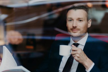 Attractive businessman in formal suit, drinks coffee during dinner break, sits in luxury cafe, looks thoughtfully at window, contemplates about business affairs has pleasant look. People and lifestyle