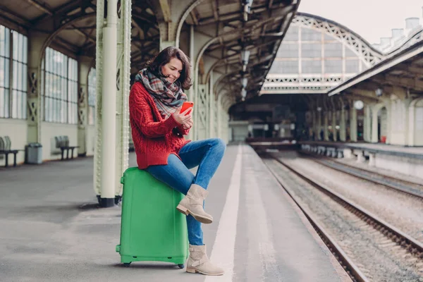 Pleased young woman has positive look, sits crossed legs at bag, holds cell phone, searches place on online map, makes video call, surfes internet website, waits for train at railway station
