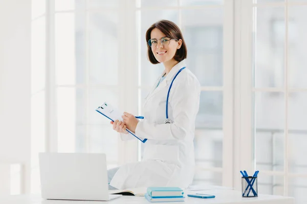 Woman doctor sits on table, writes down information in clipboard, poses at workplace in hospital office, works with laptop computer, smiles pleasantly. Health, technology, medicine, internet concept