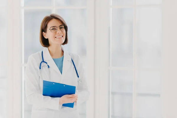 Experienced woman pediatrician stands with clipboard in cabinet, wears white medical coat with stethoscope, gives excellent medical treatment, has happy expression, ready to give consultancy