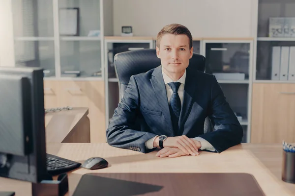 Proud handsome male entrepreneur sits in his cozy cabinet, works on computer, satisfied with successful working day, dressed formally, looks directly into camera. Intelligent employee in office
