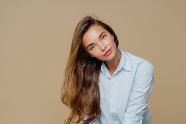 Hoş görünümlü kadın portresi sağa doğru eğik, uzun, bakımlı saçları var, mavi gömlek giyiyor, alımlı görünüme, makyaja sahip, kahverengi arka planda izole edilmiş, bilgi için boş bir alan var.