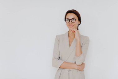 Elegant brunette businesswoman stands thoughtfully indoor, wears formal clothes, big spectacles, keeps hand under chin, stands isolated on white background, copy space for your promotion aside