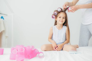 Sevimli küçük kızının yatay atış curlers rüzgarlar, beyaz arka planı poz, anaokulunda Festival olay için hazırlar annesi yakınındaki oturur. Güzel mutlu çocuk uzun saçlı