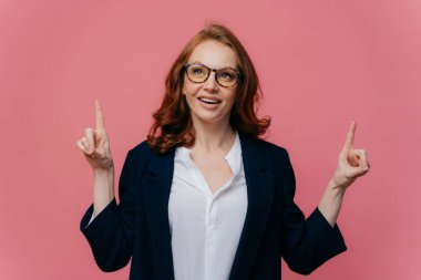 Neşeli redhaired kadın Fotoğraf yukarıda görünüyor, noktaları ön parmakyukarı bakar, yüz ifadesi memnun, bir şey gösterir, gözlük giyer, resmi takım elbise, pembe arka plan üzerinde izole