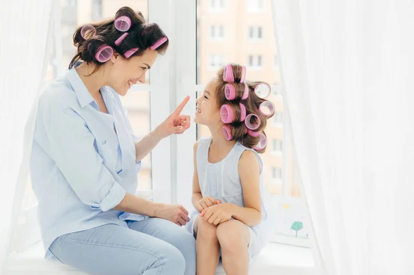 Cheerful two mother and daughter do hair nd have fun together, have curlers on head, sit on window sill. Affectionate mum touches nose of her small kid, prepare for party, care of hair and beauty