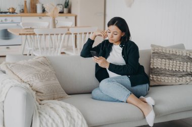 Modern female uses mobile apps at home spending leisure time online with smartphone sitting on couch. Focused young woman answers messages in social networks, browsing news, shopping on internet.