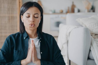 Female breathing deep, listening to meditation in earphones with namaste gesture at home. Calm woman meditates after hard day, listens to relaxing music. Stress relief, healthy lifestyle concept.