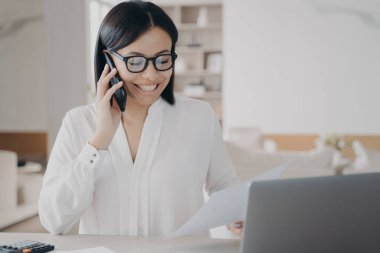 Smiling businesswoman talking on phone, answering call, works with documents, sitting at laptop. Happy female manager in glasses consulting client or enjoying corporate mobile conversation in office.