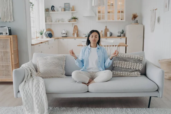 Calm female practices yoga, breathing, relaxing on modern sofa at home. Young woman meditates, sitting on comfortable couch in cozy interior. Healthy lifestyle, emotion management, stress relief.
