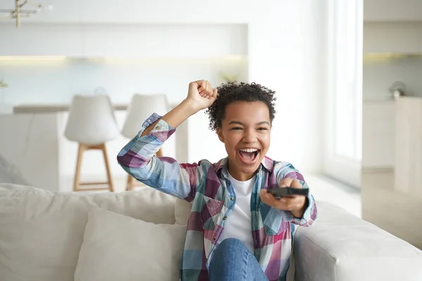 Excited happy mixed race young girl watching football on tv, screaming, makes yes winner gesture. Energetic young teen lady sport fan celebrates favorite team victory, sitting on couch at home.