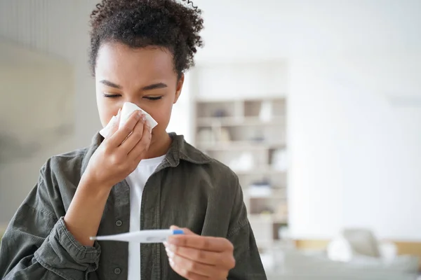 Unhealthy mixed race young teen girl blowing nose in napkin, measure temperature, using electronic thermometer, suffer from influenza flu or cold fever, has corona virus covid symptoms.