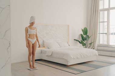 İç çamaşırlı, sabah duşundan sonra başına havluyla sarılmış sağlıklı genç bir kadın. Terazinin üzerinde dikilirken, evdeki modern yatak odasında kilo vermiş mi diye bakıyor.