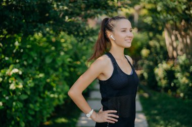 Half length shot of sporty young woman dressed in sportswear keeps hands on waist has dark hair combed in pony tail focused into distance with happy smile on face listens music poses outdoors