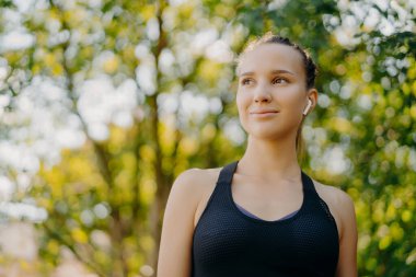 Aktif ve sağlıklı kalın. Hoş, koyu renk saçlı, spor kıyafetli bir kadın yeşil parkta durup, kulaklıkla en sevdiği müziği dinledikten sonra mutlu bir ifadeyle bakışlarını kaçırıyor.