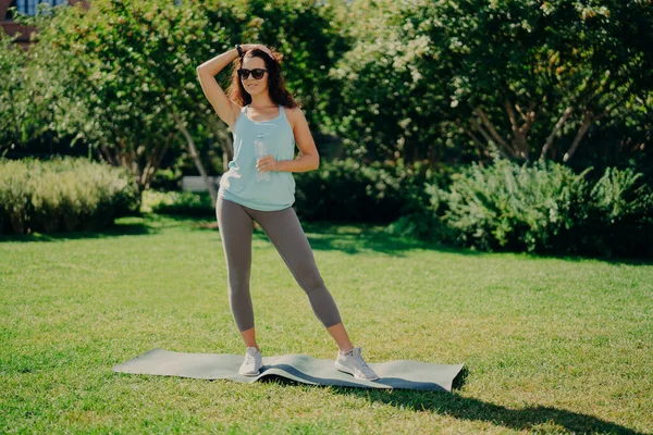 Fitness sport and recreation concept. Full length shot of sporty woman dressed in t shirt leggings and sneakers stands on karemat holds bottle of water poses against green nature background.