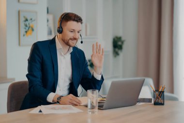 Smiling young businessman in headset and suit saying hello, waving to somebody and smiling while working remotely at home, having video call with colleagues or taking part in web conference