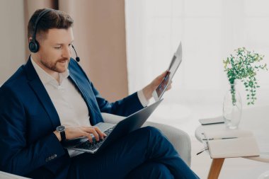 Sorumlu, takım elbiseli ve kulaklıklı bir erkek çalışanın elinde evrak ve dizüstü bilgisayarla oturma odasında otururken ve online toplantı yaparken uzaktan evde çalışan odaklanmış bir işadamı.