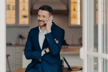 Resmi takım elbiseli genç ve çekici bir işadamı telefonda konuşuyor ve iyi haberler duyduğunda gülümsüyor oturma odasında masaya yaslanıyor, akıllı telefon tutuyor ve proje sonuçlarını tartışıyor. İş konsepti