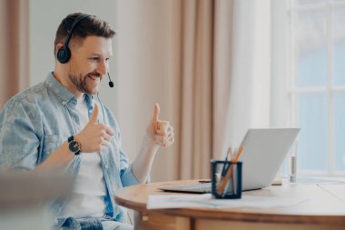 Tatmin olmuş sakallı erkek model evde online eğitime katılıyor. Kulaklık kullanıyor. İki eliyle diz üstü bilgisayara dikkatlice bakıyor.