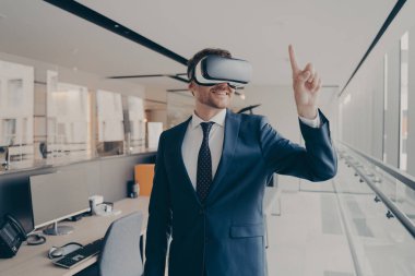 Siber uzay tecrübesi. VR gözlük takan mutlu ofis çalışanı dijital dünyadaki objeleri işaret ediyor, üç boyutlu gözlüklü iş adamı sanal gerçeklikle etkileşimde, takım elbise giymiş.