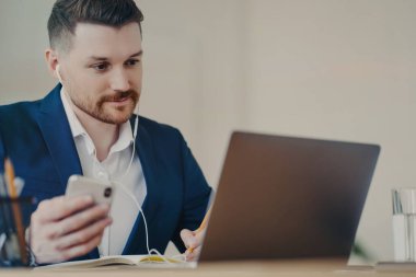 Odaklanmış erkek yönetmen kablosuz kulaklıklarla kurumsal giysiler giyip cep telefonuyla dizüstü bilgisayar başında iş webinar 'ı izliyor ya da rahat bir dolapta video görüşmesi yapıyor. İş mesleği kavramı
