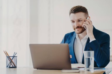 Genç yakışıklı iş adamı dizüstü bilgisayarın önünde telefonda konuşurken dikkatle dinliyor ve laptoptan bilgileri kontrol ediyor, şık bir ofiste minimal tasarım ile oturuyor.