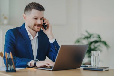 Resmi takım elbiseli başarılı bir girişimci, cep telefonuyla konuşuyor, iş ortaklarıyla iş görüşmeleri yapıyor, modern ofisteki çalışma masasının arkasında modern laptopun önünde oturuyor.