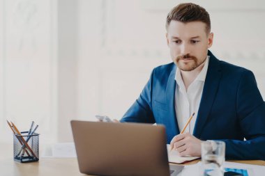 Çalışma sürecinde yer alan ciddi sakallı pazarlamacı kalemle not alır. Bilgisayara dikkatlice bakar, mavi takım elbise giyer, ortak çalışma alanı düzenleme planı yazar.
