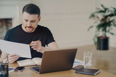 Yakışıklı, serbest çalışan erkek, siyah tişörtlü ya da işadamı, evden uzaktan çalışırken bilgi ya da satış raporlarıyla belgeyi analiz ediyor, dizüstü bilgisayarla ve diğer ofislerle aynı masada oturuyor.