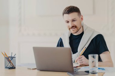 Kulaklıkla gülümseyen genç bir adam meslektaşlarıyla video görüşmesi sırasında notlar yazıyor ya da internetteki iş webinar 'ı, ana ofiste otururken dizüstü bilgisayar kullanıyor, evden uzaktan çalışıyor.