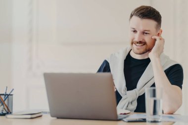 Sıradan kıyafetler giyen, kulaklık takan, online toplantı yapan iş adamı, ofis masasının arkasında dizüstü bilgisayarın önünde oturmuş iş arkadaşlarıyla sohbet eden pozitif erkek.