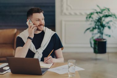 Evden çalışan mutlu genç bir işadamı, iş yerinde telefonla konuşan, gülümseyen, dizüstü bilgisayarı ve bir bardak suyu olan serbest çalışan bir erkek. Uzak görev konsepti