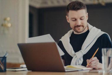 Proje stratejisi üzerinde çalışan günlük kıyafetlerle odaklanmış işadamı, iş fikirlerini deftere yazıyor, evdeki veya ofisteki modern dizüstü bilgisayarla masada otururken belge tutuyor. İş konsepti
