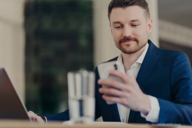 Zarif kıyafetler giymiş ciddi erkek girişimcilerin görüntüsü modern cep telefonu ve dizüstü bilgisayar ile yeni projeler üzerinde çalışıyor. Çağdaş ofiste bulanık arka plana karşı kapalı pozlar veriyor..