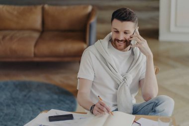Neşeli, serbest çalışan genç bir adam ya da erkek girişimci cep telefonuyla müşteriyle konuşuyor ve uzaktan çalışırken not defterine bazı notlar yazıyor, oturma odasında deri koltukla oturuyor.