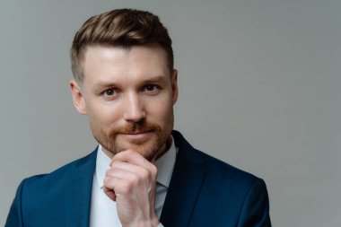 İş adamları. Mavi takım elbiseli yakışıklı, tıraşsız iş adamı veya erkek girişimcinin portresi çenesine dokunuyor ve gri stüdyo geçmişine karşı dikkatlice kameraya bakıyor.