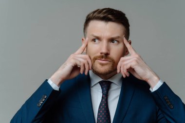 Let me think. Thoughtful unshaved businessman or male entrepreneur in suit holding fingers on his temples and looking aside while posing against grey studio background, headshot portrait