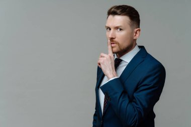 Keep it in secret. Young businessman in formal wear suit showing shh sign, keeping finger near mouth while standing against grey studio background, looking at camera with shushing expression